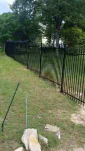 An ornate metal driveway gate with stone pillars and a matching wooden rail fence installed by Ft Worth Fence And Patio in Fort Worth, TX.