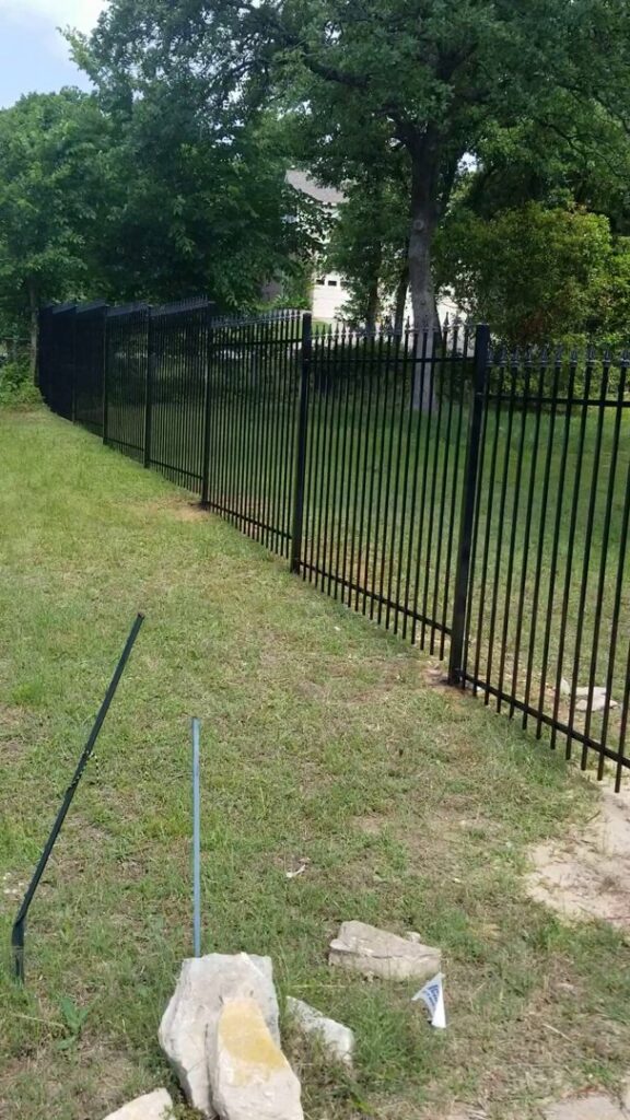 An ornate metal driveway gate with stone pillars and a matching wooden rail fence installed by Ft Worth Fence And Patio in Fort Worth, TX.
