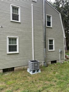 An outdoor AC condenser unit with a metal flue pipe installed next to a house by Foremost Mechanical Services in Hartford, CT.