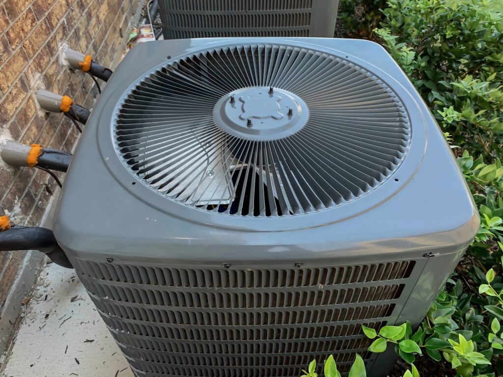An outdoor air conditioning condenser unit situated next to a brick wall and green bushes, installed by Evergreen HVAC Comfort Solutions in Milwaukee, WI.