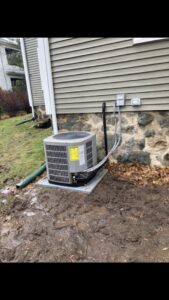 An outdoor air conditioning unit installed next to a house in a muddy area by Freedom Air Mechanical Contractors LLC in Everett, MA.
