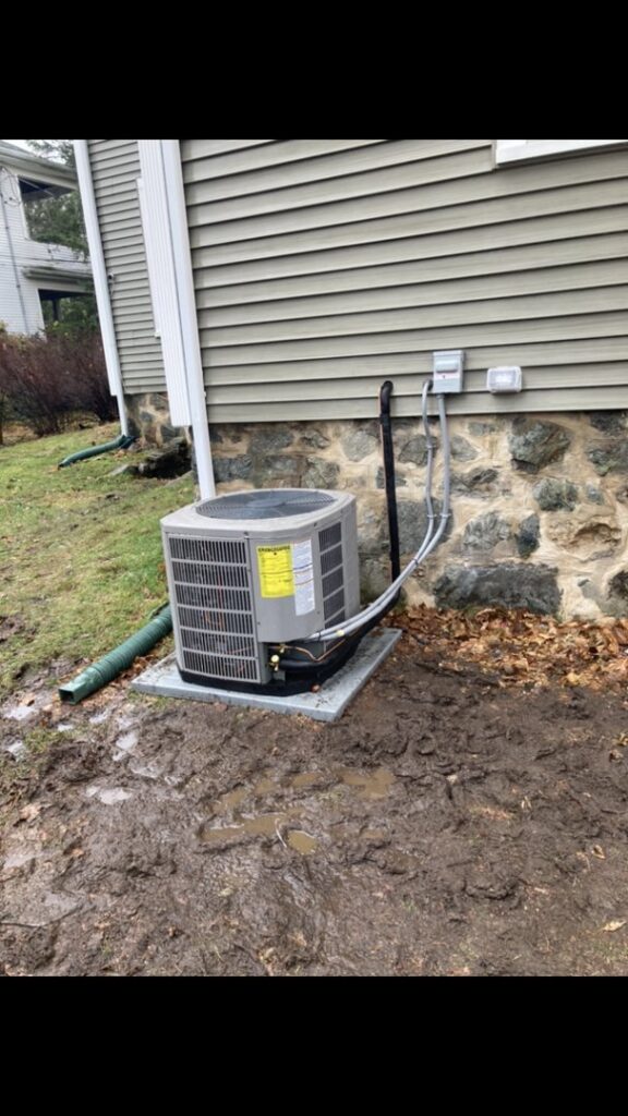 An outdoor air conditioning unit installed next to a house in a muddy area by Freedom Air Mechanical Contractors LLC in Everett, MA.