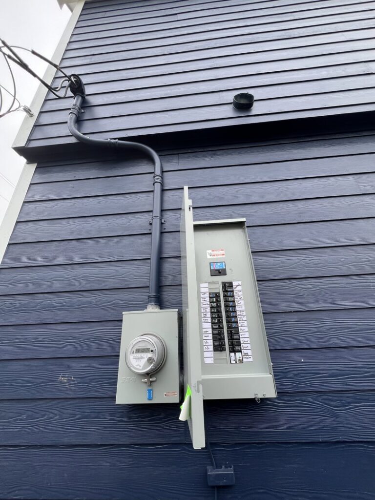 An outdoor electrical meter and main breaker panel installed on a blue siding wall by Llanos Electric Builders in Houston, TX.