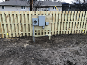 An outdoor electrical meter and service panel installed on a wooden fence by Hills Electric in Rowley, MA.