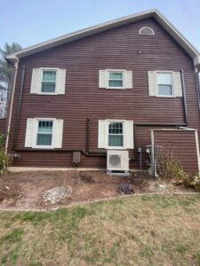 An outdoor heat pump unit installed next to a brown house by Champlain Valley Heat Pump, LLC in Colchester, VT.