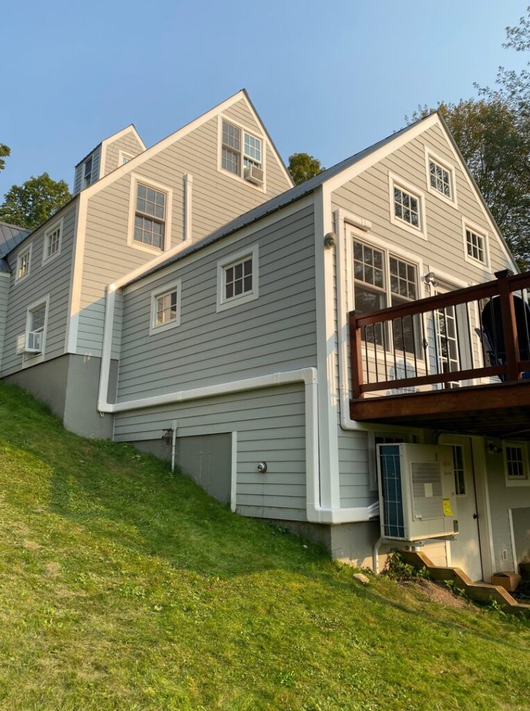 An outdoor heat pump unit installed on the side of a gray house by Champlain Valley Heat Pump, LLC in Colchester, VT.