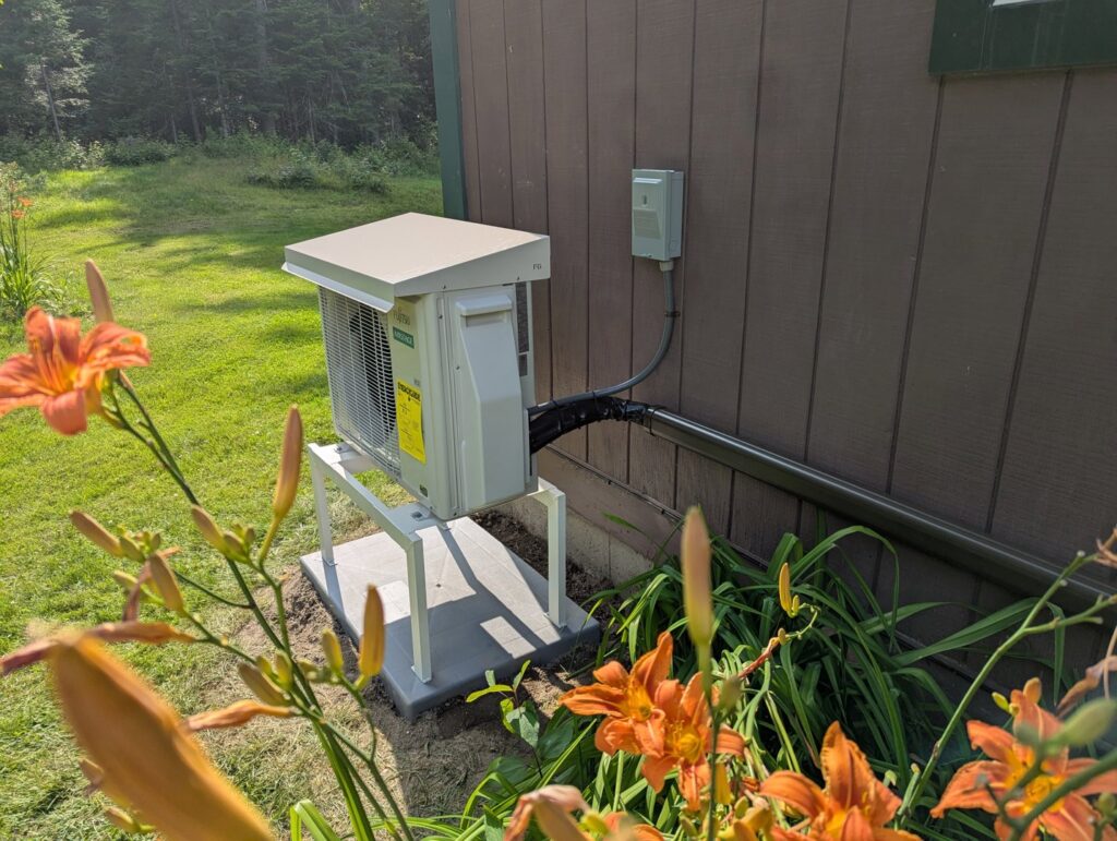 An outdoor heat pump unit installed on the side of a house with landscaping by Kinney Family Heat Pumps in Augusta, ME.