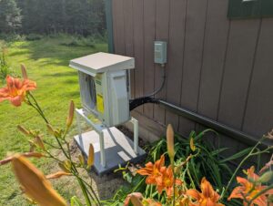An outdoor heat pump unit installed on the side of a house with landscaping by Kinney Family Heat Pumps in Augusta, ME.