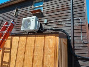 An outdoor HVAC condenser unit installed on the side of a wooden building, with an orange ladder, by Jones Heating and Refrigeration in Dickinson, ND.