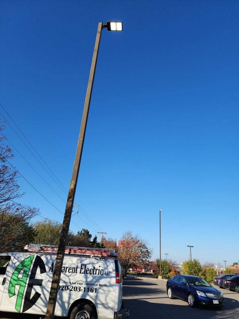 An Apparent Electric LLC service van parked next to a tall outdoor light pole, indicating commercial lighting service in Oshkosh, WI.