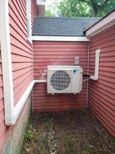 An outdoor mini-split air conditioning unit professionally installed on the side of a red house by Shook Heating And Cooling in Livonia, MI.