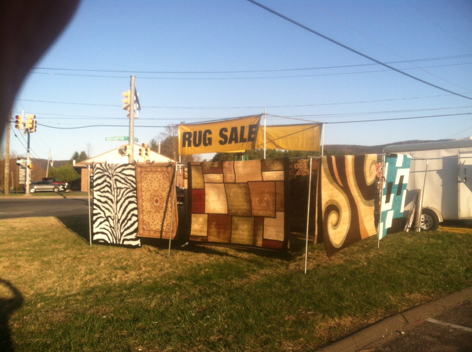An outdoor display of various rugs for sale under a 'RUG SALE' banner by Sunshine Rugs and Art in Londonderry, NH.