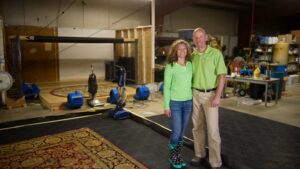 The owners standing in their workshop surrounded by professional carpet cleaning equipment at All Seasons Carpet Cleaning in Loves Park, IL.