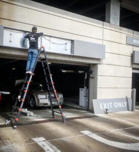 An electrician from Lumenus Technologies, LLC repairing an overhead light in a parking garage in Lenexa, KS.