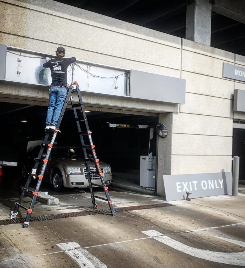 An electrician from Lumenus Technologies, LLC repairing an overhead light in a parking garage in Lenexa, KS.