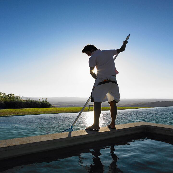 A person in silhouette using a skimmer to clean an infinity pool, showcasing Bryan Simpson's pool services in Services Available In.