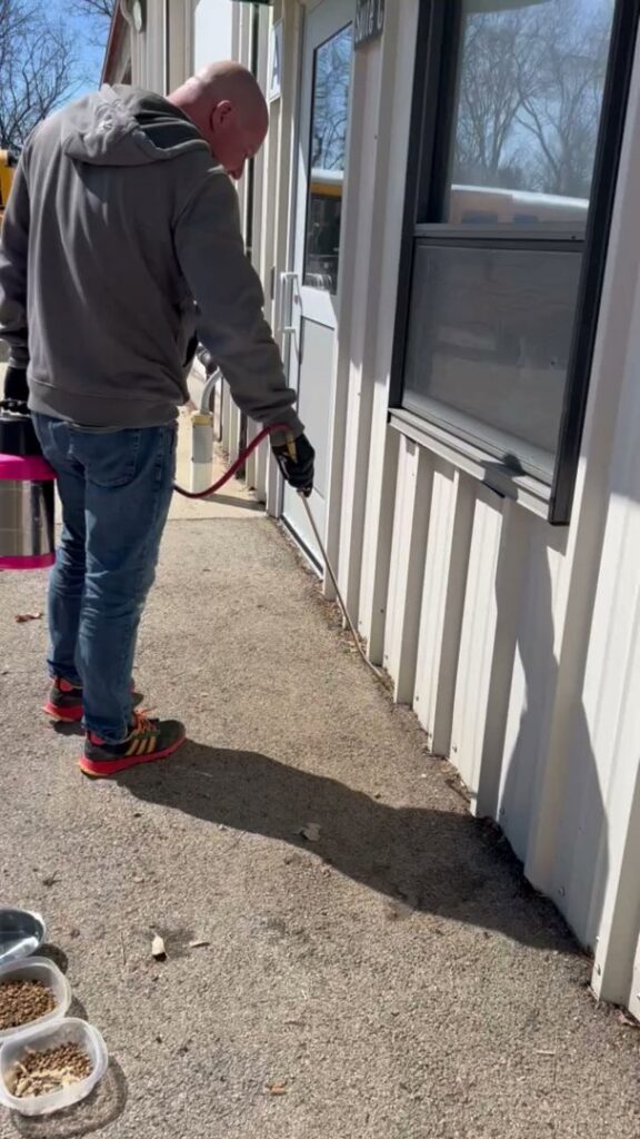 A pest control expert spraying along the foundation of a commercial building for Modern Solutions Pest Control in Rockford, IL.