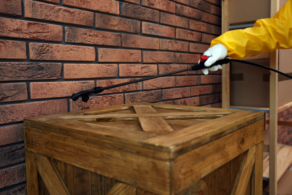 A hand in a yellow glove spraying a brick wall above a wooden crate for pest control by Venum Pest Solutions in Fayetteville, NC.