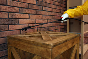A hand in a yellow glove spraying a brick wall above a wooden crate for pest control by Venum Pest Solutions in Fayetteville, NC.