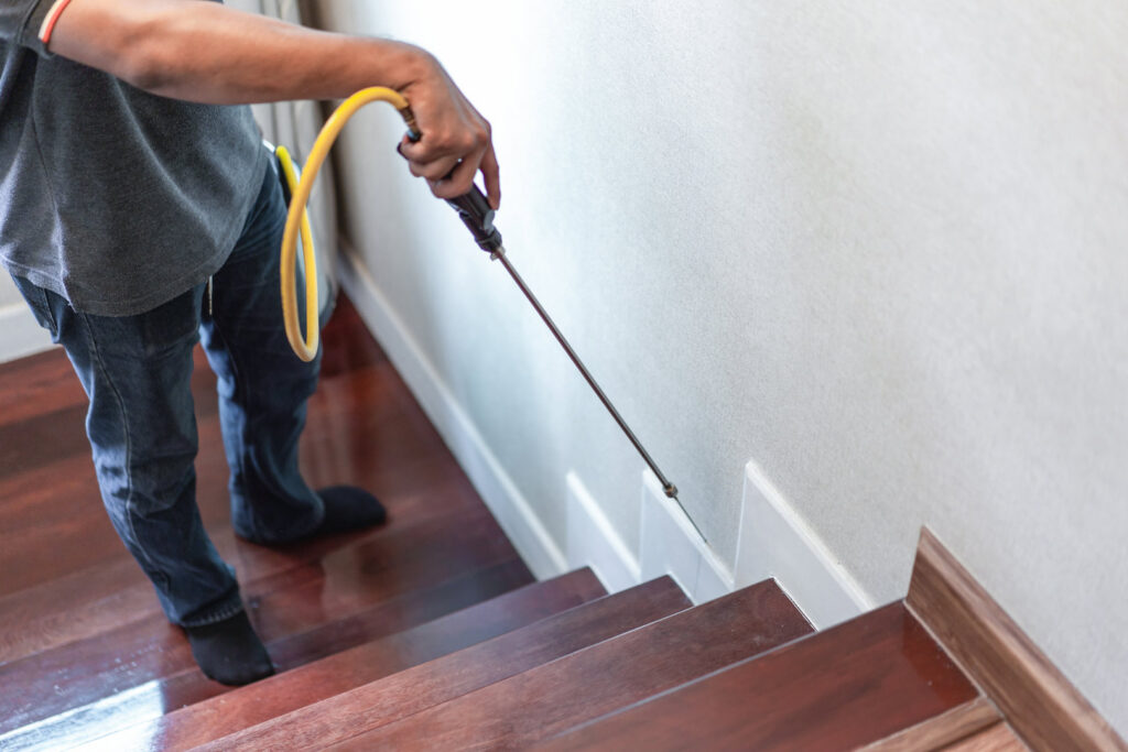 A pest control technician spraying along the baseboards of a wooden staircase to eliminate pests for Ares Pest Control in Biddeford, ME.