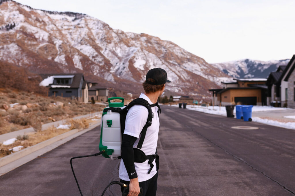 A Peak Pest Control technician wearing a backpack sprayer on a residential street in Ogden, UT, ready for service.