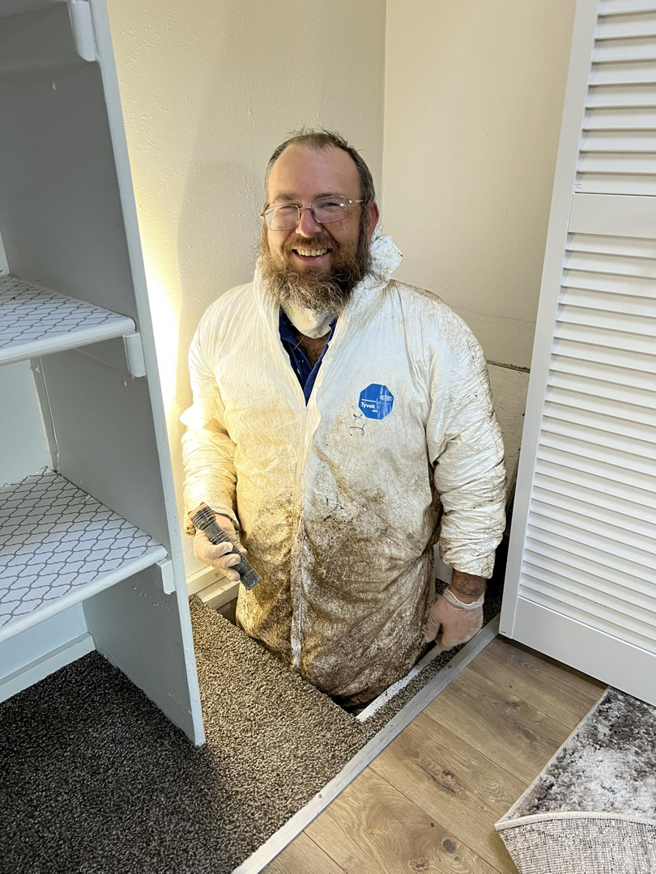 A pest control technician in a protective suit emerging from a crawl space at a job site for Top Gun Pest Control in Caldwell, ID.