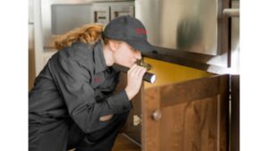 A pest control technician inspecting under a kitchen cabinet with a flashlight for X-Out Pests Control in Little Rock, AR.