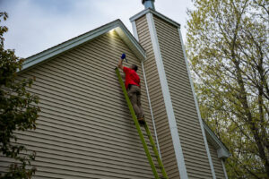 A pest control technician on a ladder inspecting a house exterior for pest control at Big Blue Bug Solutions in Providence, RI