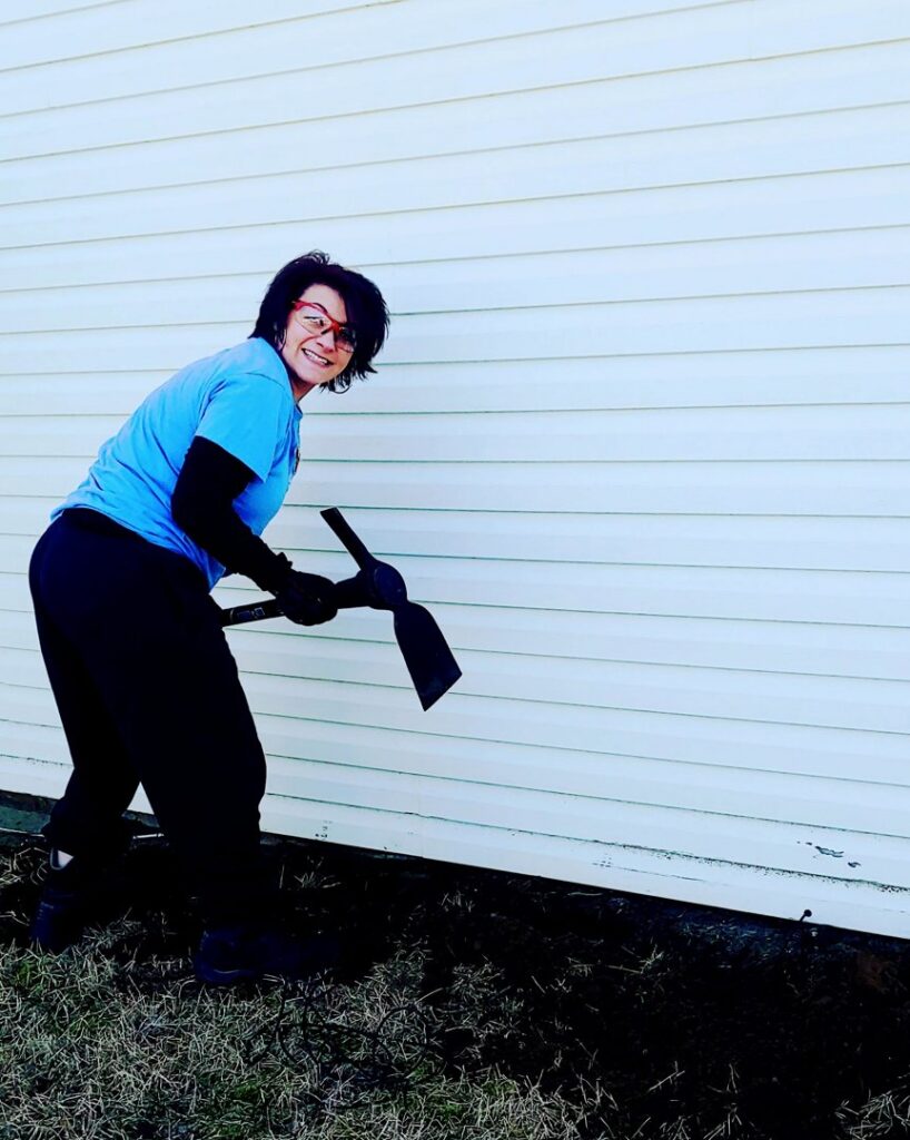 A Mario's Pest Control technician using a tool to prepare the ground for a pest treatment around a house foundation in Hamilton, NJ.
