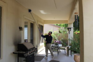 A pest control technician removing cobwebs from a patio ceiling with a long duster by Overson Pest Control LLC in Mesa, AZ