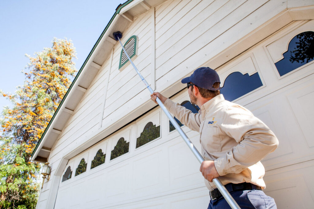 A Crazy Ant Pest Control technician removing cobwebs from a house exterior with a long pole in Fresno, CA.