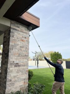 A CAPE Pest Control technician removing cobwebs and nests from house eaves with a long pole in Mesa, AZ