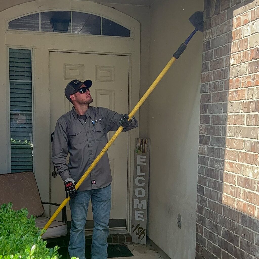 A pest control technician from Kaizen Pest Management removing spiderwebs from a house exterior in Austin, TX.