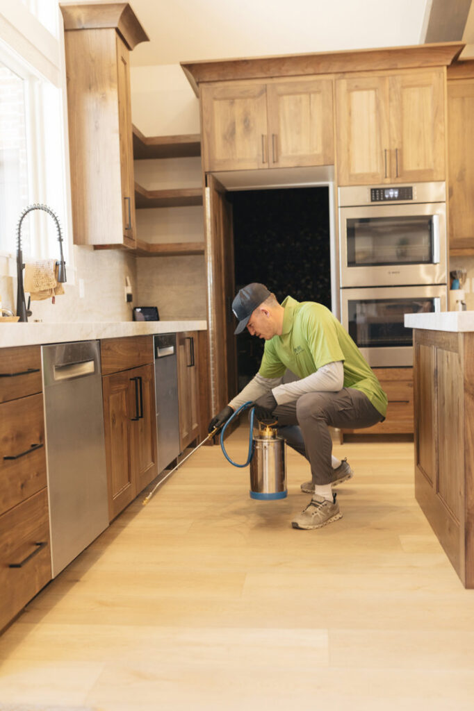 A pest control technician spraying along kitchen cabinets for interior pest treatment at Pure Pest Solutions in North Ogden, UT.