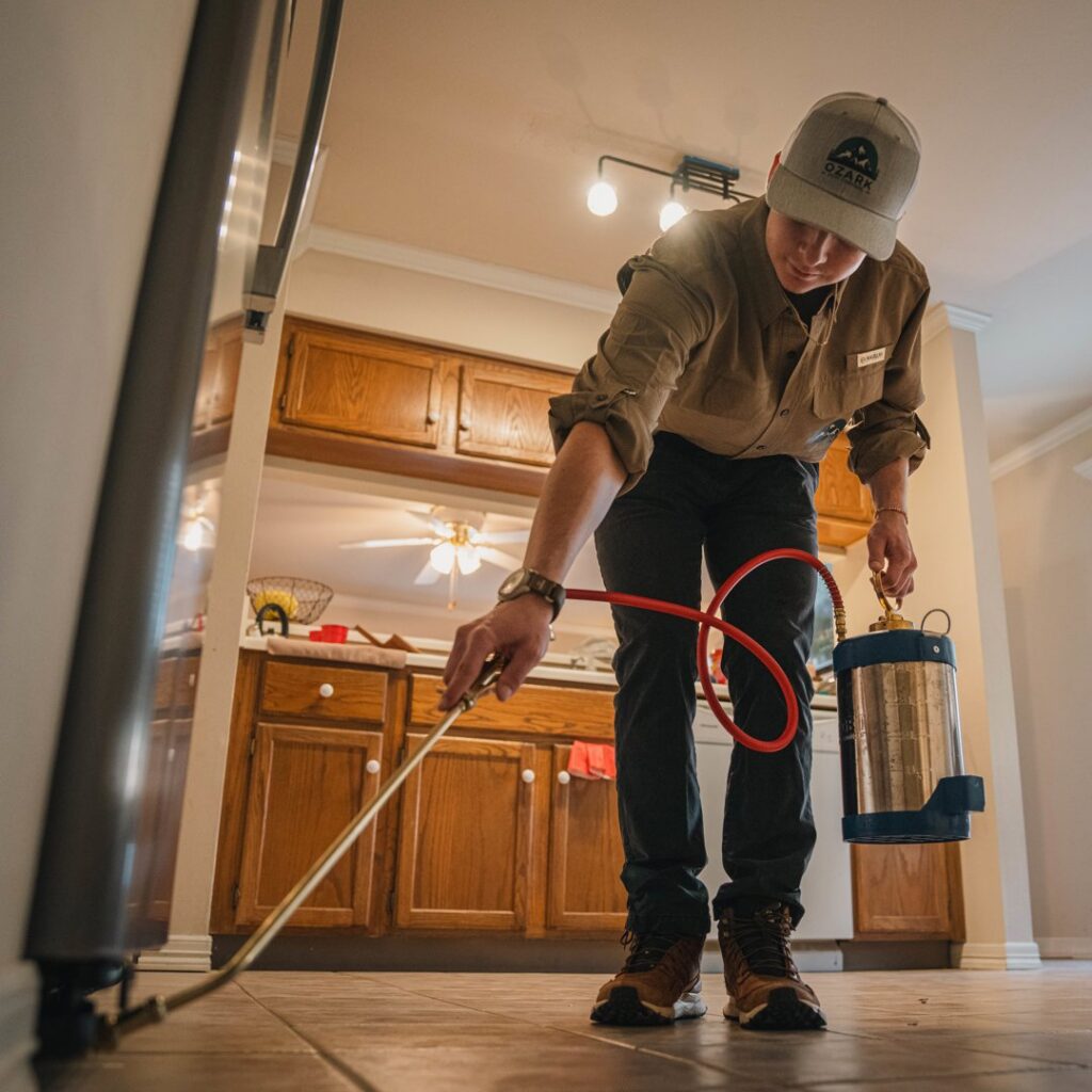 A pest control technician spraying along kitchen baseboards during an interior treatment by Ozark Pest Control in Fayetteville, AR.