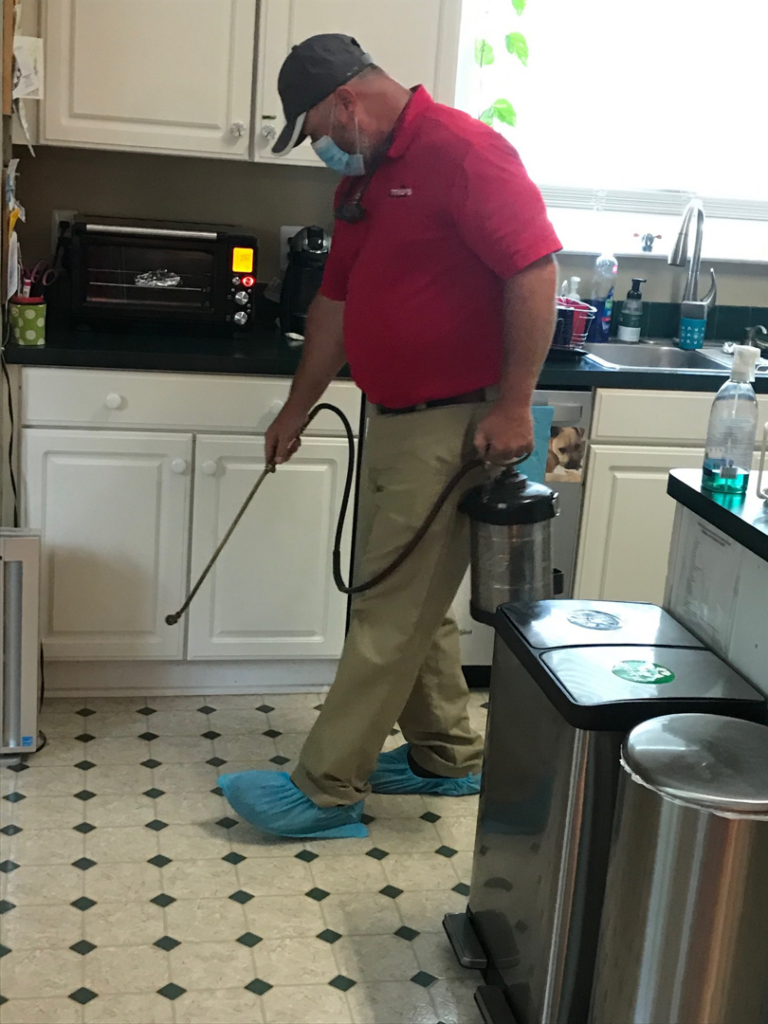 A pest control technician wearing a mask and shoe covers, spraying for pests inside a kitchen at Otho's Pest Management in Randleman, NC