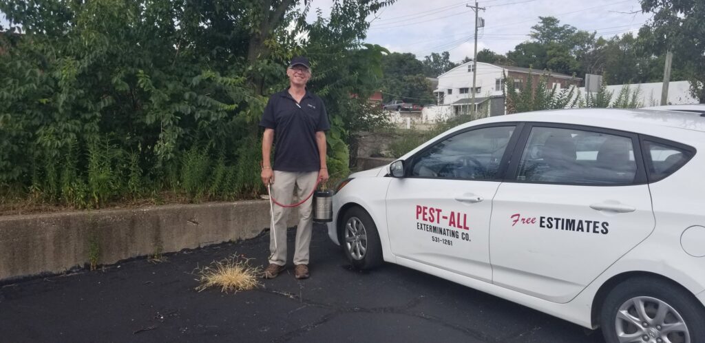 A pest control technician holding a sprayer next to a company car for Pest-All Exterminating Co. in Cincinnati, OH.