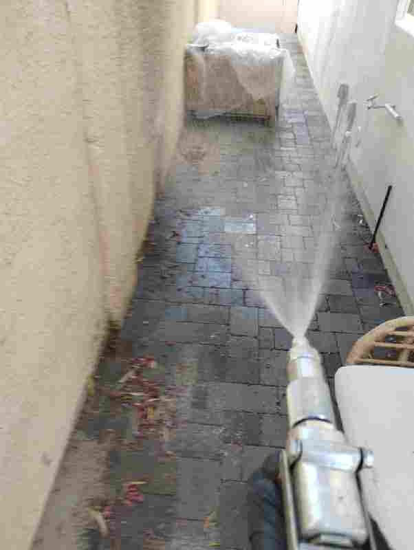 A pest control technician spraying pesticide along a paved walkway next to a building wall, providing service for Bernard's Pest in Glendale, AZ.