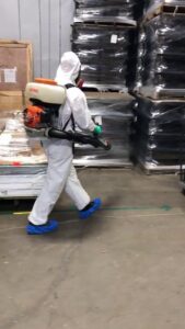 A pest control technician in a hazmat suit using a backpack sprayer in a warehouse for Ameri Care Services, Inc. in Murfreesboro, TN.