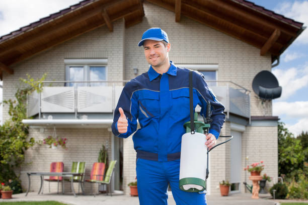 A smiling pest control technician giving a thumbs-up with a sprayer in front of a house for Grenier's Pest Control in Essex Junction, VT.