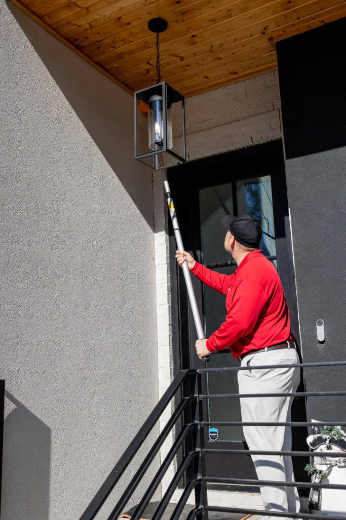 A pest control technician treating an exterior light fixture or eave with a long pole for DOA Pest Service in Nashville, TN.