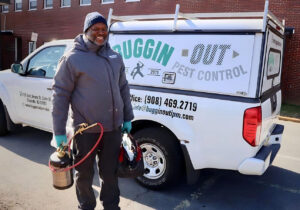A pest control technician holding a sprayer and helmet next to a branded truck for Buggin Out Pest Control in Elizabeth, NJ
