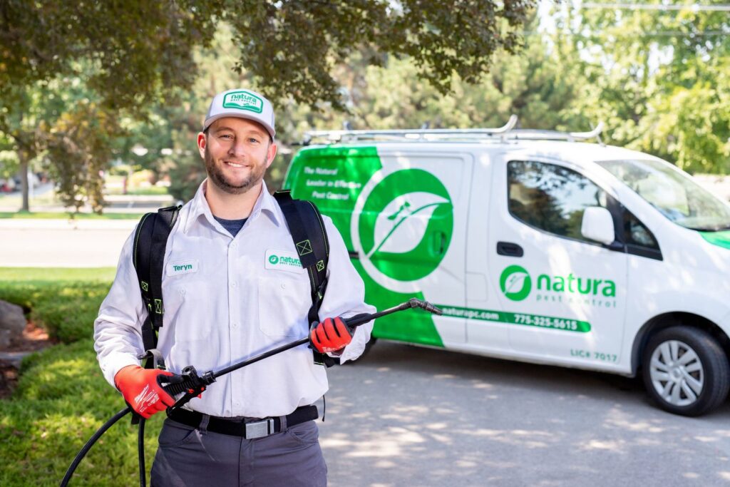 A Natura Pest Control technician wearing a sprayer backpack stands in front of a branded van in Boise, ID.