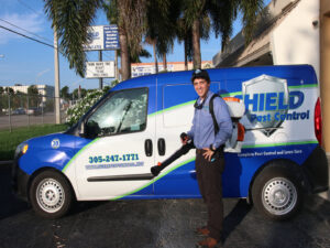 A Shield Pest Control technician holding a pest control sprayer next to a branded service van in Homestead, FL.