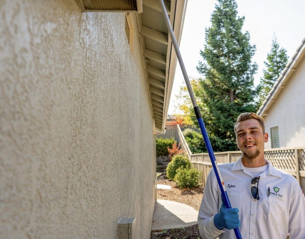 A smiling pest control technician from The Noble Way Pest Control holding a web removal brush at a home in Sacramento, CA.