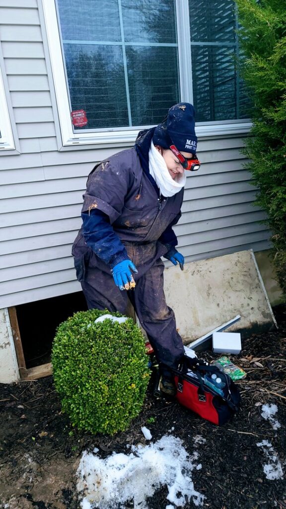 A Mario's Pest Control technician in protective gear working near a crawl space entrance for pest control in Hamilton, NJ.