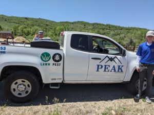 Two Peak Pest Control technicians standing next to a branded truck with a 'Lawn Pest' logo in Ogden, UT.