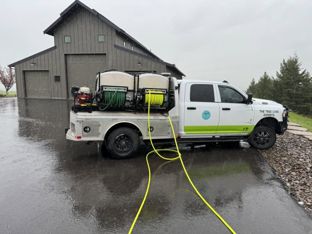 A Pest One service truck with hoses extended, performing pest control or plant health treatment on a driveway in Bozeman, MT.