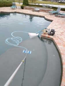 A pool cleaner actively brushing the bottom of a murky pool with a long pole, with a pump on the deck, by Brian's Pool Service in Baton Rouge, LA.