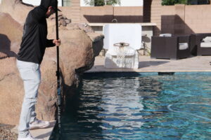A pool technician actively skimming or cleaning a swimming pool with a long pole, performed by Pool Rescuers LLC in Gilbert, AZ.
